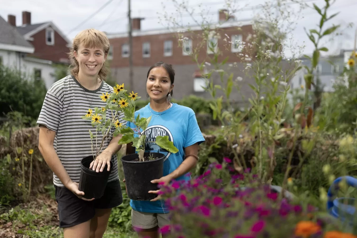 Two students stand together in a garden, surrounded by plants, while holding potted plants and smiling