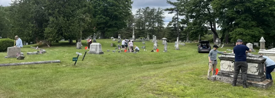 People in a graveyard cleaning stones. On the right a group of three people work on a large marble stone scraping off moss and lichen. On the left a woman rakes leaves and grass clippings away from a stone. In the center a group of people scrapes lichen and moss from a marble pillar. 