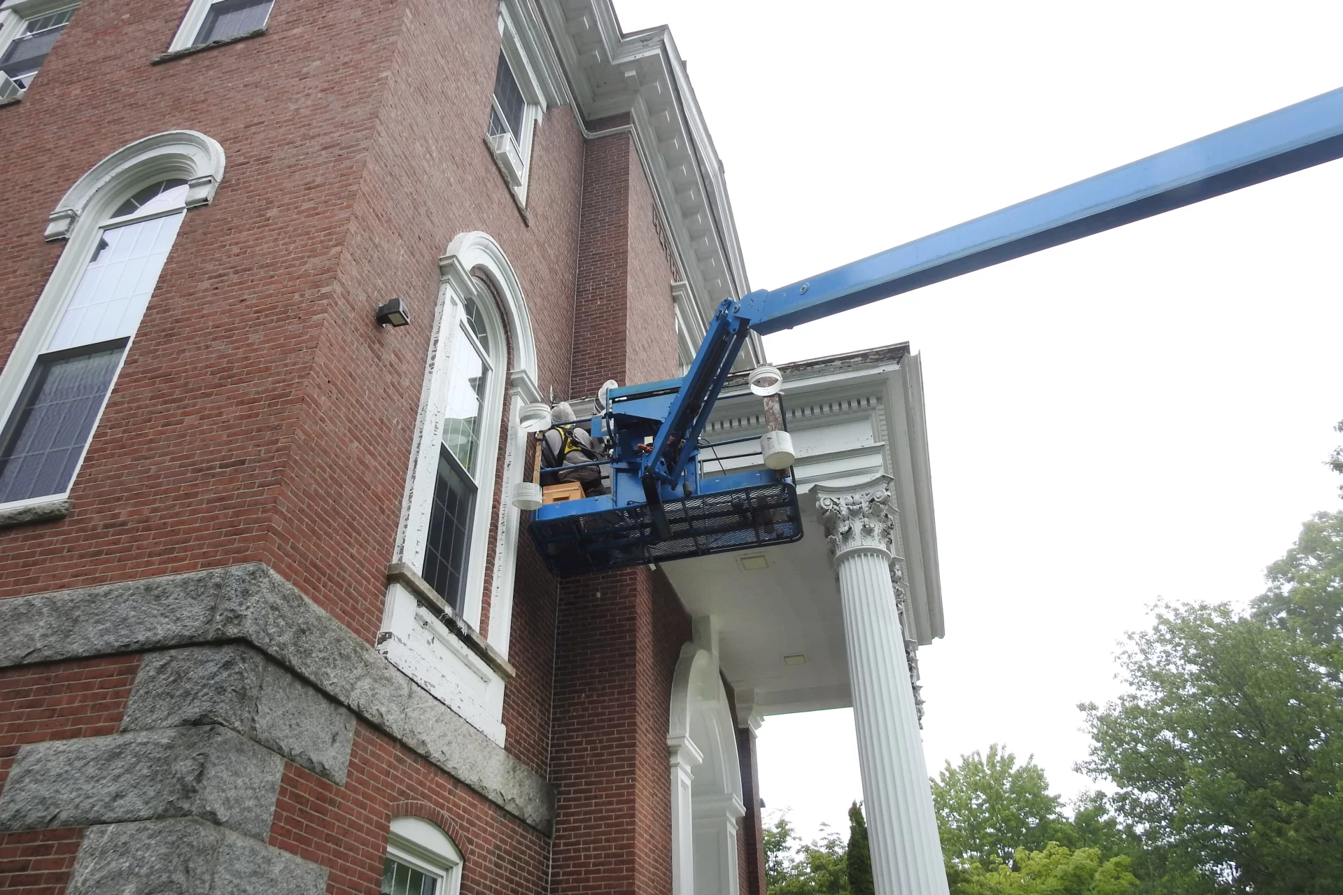 Brian Mason of Spicer Bees (purple shirt) removes a big honeybee hive from the college's oldest building, Hathorn Hall, on July 25, 2024.

Discovered by a work crew painting the Hathorn trim, the hive was in a section of the soffit of the Hathorn portico, where the roof joins the main part of the building. 

Lifted to the second-story location by a Genie boom lift, Mason and Bates carpenter Matt Capone, with beard, worked together, the latter using a multitool and pry tools to remove trim to get at the hive, and the former using a homemade vacuum and hive tools to remove the bees and comb, relocating as much of the hive as possible to his apiary in Whitefield. 

Mason, who estimated the hive at upwards of 50,000 bees, gives credit to Bates for calling in a beekeeper for the project. "100 percent credit," he says, noting that the Bates hive was not only healthy but had apparently survived for at least one winter, if not more. Most wild honeybee hives die each winter, and commercially maintained hives typically rely on treatments to control mites.

"Less human intervention is always valuable. The fact that this hive wintered without intervention means the bees might just have something going for them to survive. So now I can introduce the Bates bees into my apiary. That’s a win."