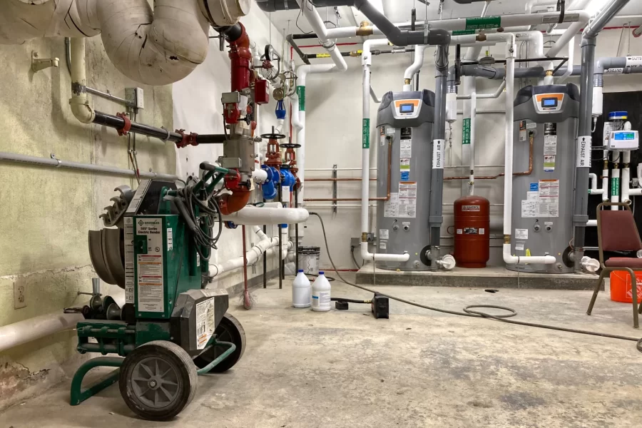 The mechanical room at 96 Campus Avenue. From left, features include sanitary discharge, water intakes for fire suppression and domestic water, and two gray heaters for domestic hot water. The wheeled contraption is used for bending electrical conduits. (Doug Hubley/Bates College)