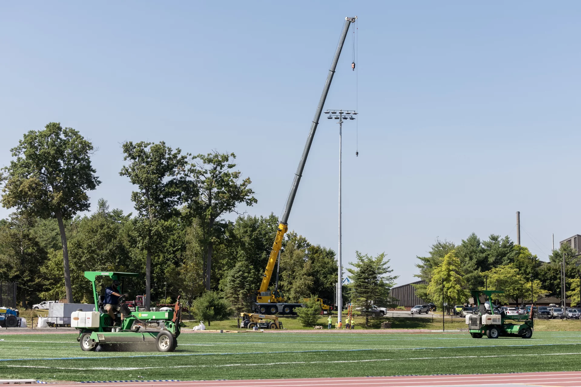 A Cote Corp. crane has just finished placing a new field light, one of four, at Russell Street Field on Sept. 5. Meanwhile, two machines groom the crumb rubber and sand infill on the field’s new playing surface. (Theophil Syslo | Bates College)