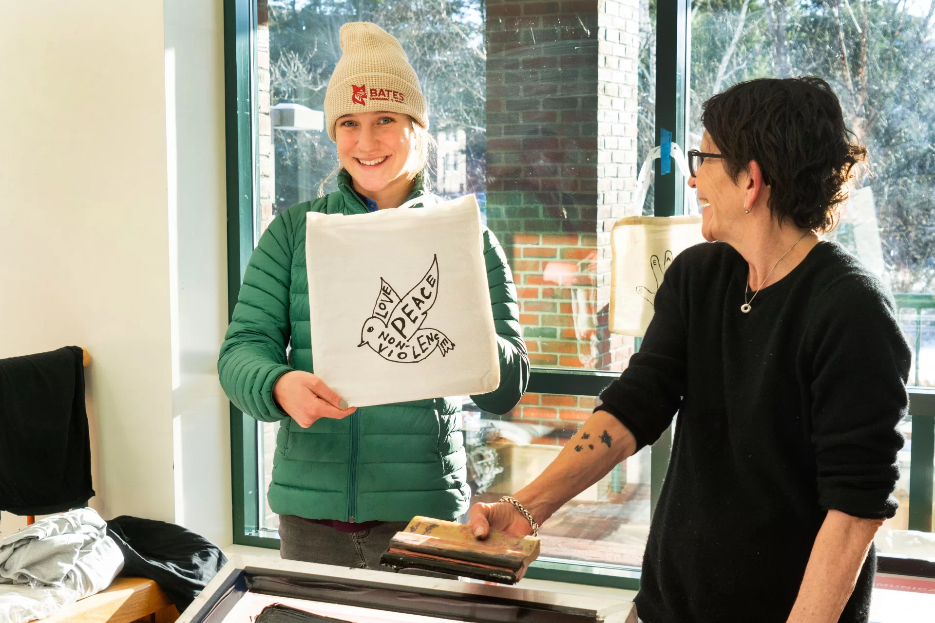 January 20, 2025 - January 20, 2025 - Professor Michel Droge(in black) and Professor Cat Balco (in pink) lead a Screen Printing Drop-In Workshop relating to the college’s 2025 MLK Day theme, Bending Toward Justice: Peace and Nonviolence. T-Shirts and Tote bags were available for attendees to screen print one of the student created designs. 

Sarah Van Lonkhuyzen ‘27 (left) from Rockport, Maine was one of the first students to ink up the themed screen prints.